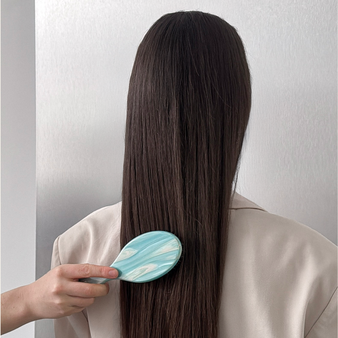 A person viewed from the back with long, straight dark brown hair, using a Butr Hair Essentials light blue and white marbled oval hairbrush. The person is wearing a beige top against a plain light gray wall.