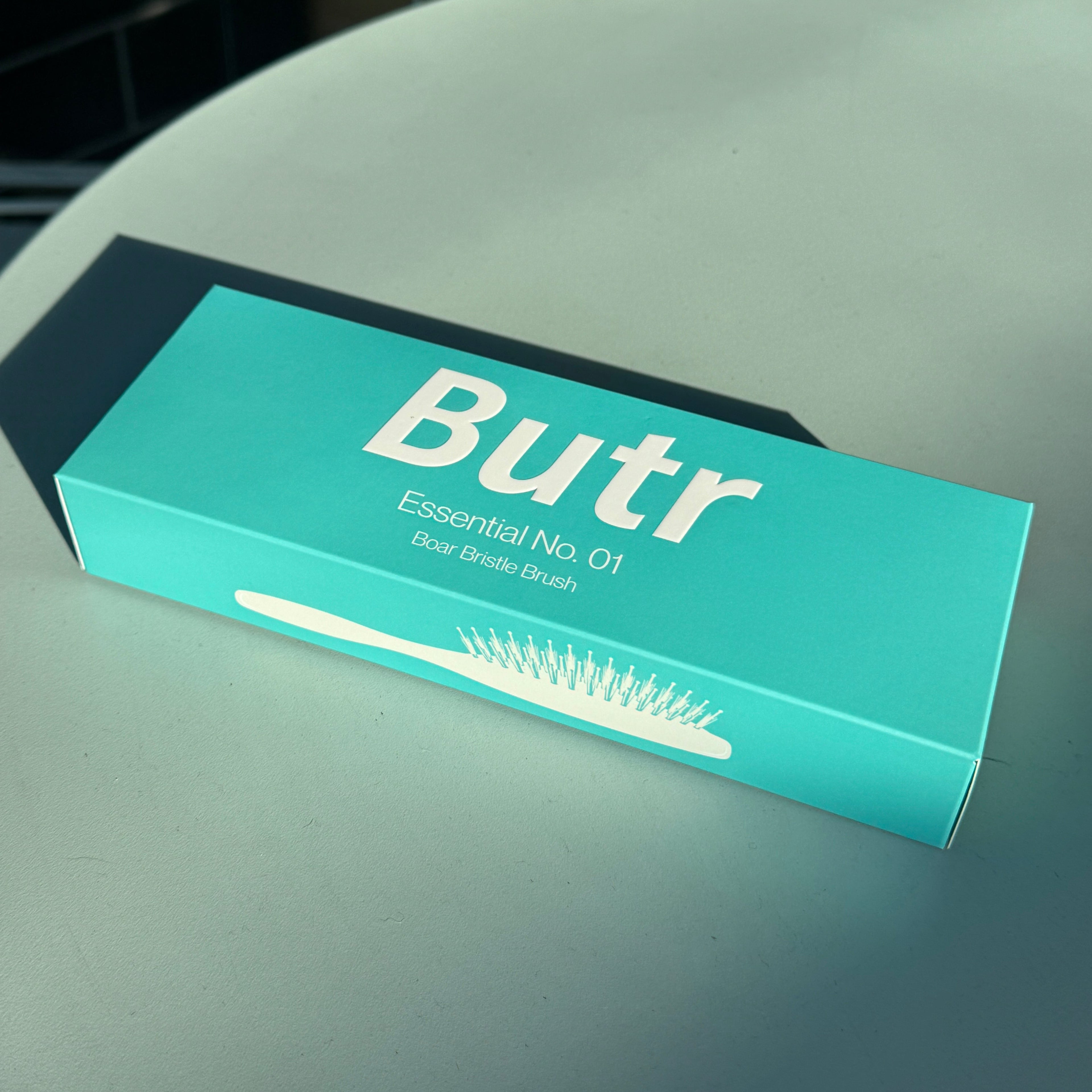 Butr Hair Essentials teal and rectangular box for the Butr Essential No. 01 Boar bristleBrush, photographed on a gray tabletop.